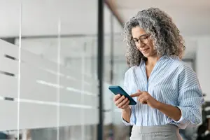 A woman in a striped shirt and glasses is looking at her phone in an office building with glass walls.