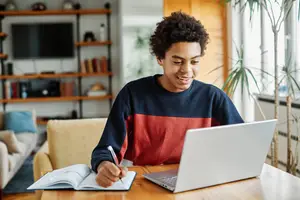 A young man is smiling and writing on a notepad while using a laptop on a wooden table in a room.