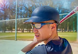 A young man is standing on a baseball field, holding a baseball bat, and wearing a blue helmet and sunglasses.