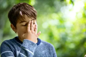 Young boy wearing a blue sweater with a white stripe on the sleeves is rubbing his face with his right hand while closing his eyes, probably feeling a headache