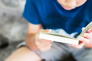 A young boy is sitting on the floor, holding a book in his hands, with his eyes focused on the book, possibly reading it.