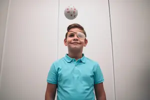 A boy wearing a blue polo shirt and a white eye patch is playing with a white ball with red letters.