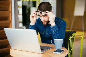 A man in a denim shirt adjusts his glasses while looking at a laptop on a table with a cup and a phone.