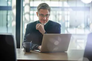 Business man in a suit adjusting glasses and smiling while sitting at a table in an office.