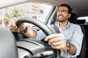 A smiling man wearing glasses and bracelets drives a car with his hands on the steering wheel.
