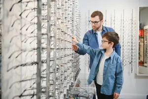 A man and a boy are standing in front of a wall with hanging eyeglasses in a shop.