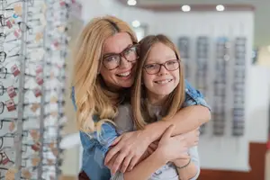 A woman with glasses is hugging a young girl with glasses in a store with glass racks in the background.