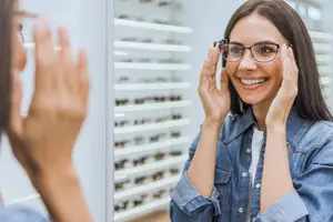 A smiling woman wearing glasses is looking at herself in a mirror while holding her glasses up to her face.