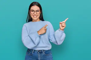 A woman wearing a blue sweater and glasses is pointing her finger at a blue background while smiling.