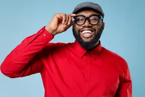 A man wearing a red shirt and glasses, smiling, and touching his glasses with his left hand, standing in front of a blue background.