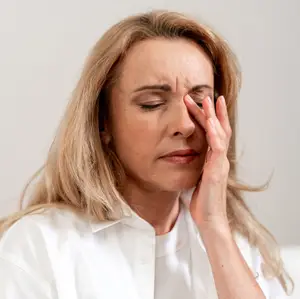 A woman with blonde hair in a white shirt is holding her forehead with her left hand and closing her eyes, looking down, and seems to be feeling pain. She is probably sitting on a couch, and her face is slightly blurry, and her hair is slightly messy.
