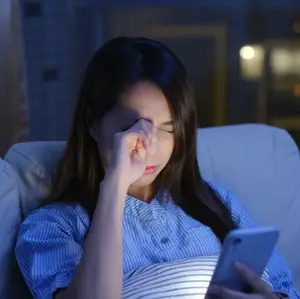 A young woman sitting on a couch and looking at her phone while holding her nose and closing her eyes.