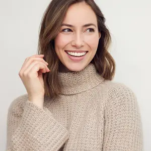 A woman wearing a beige turtleneck sweater smiles and poses for a photo.
