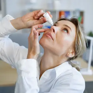 A woman in a white lab coat is administering eye drops to herself.