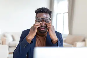 African American man wearing glasses and suit jacket sitting in front of computer screen, smiling with hand on face