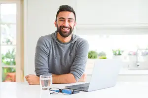 A man in a grey hoodie sits at a table in a kitchen with a laptop, a glass of water, and glasses. He has his arms crossed and is smiling.