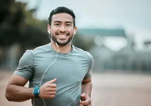 A man wearing a gray t-shirt, a watch, and earphones is jogging and smiling in a park