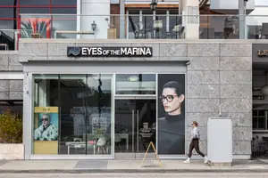 A woman walks past a storefront with the name Eyes of the Marina and a billboard featuring a woman wearing glasses