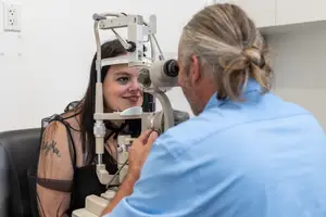 A woman with a tattoo on her left arm is getting her eye examined by an optometrist in a blue shirt.