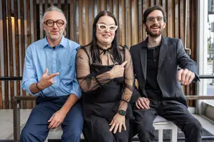 Three individuals sitting on a bench and smiling at the camera, with a wooden wall in the background