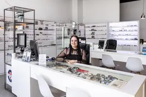A smiling woman in a black blouse sits behind a desk in a store filled with glasses