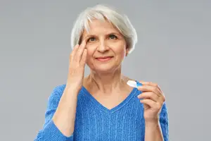 An elderly woman with gray hair and blue eyes holding a contact lens and looking at the camera with a smile