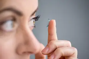 A woman is wearing a blue contact lens in her eye and is about to put it in her eye.