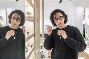 A man with curly hair is adjusting his glasses in front of a mirror while standing in a room with wooden shelves, white walls, and a plant