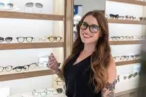 A woman wearing glasses is smiling and posing in front of a shelf of glasses.