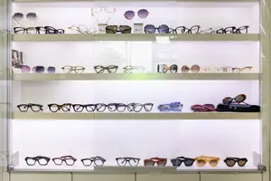 A glass display case filled with various sunglasses and eyeglasses, each pair showcased on a white background with a reflection of the white wall behind the display.