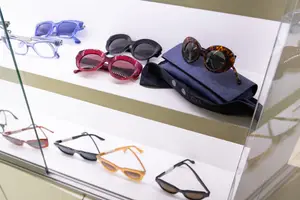 A collection of sunglasses are displayed on a white shelf in a shop, including a blue pair with a silver frame, a red pair with a red frame, a black pair with a black frame, a blue pair with a brown frame, a blue pair with a white frame, a black pair with a brown frame, and a black pair with a black frame, all protected by a glass cover.