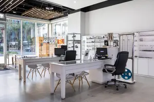 An empty interior of a business establishment with glass walls and doors, showcasing various office furniture like desks, chairs, and monitors, and a glass display case with frames, all under bright lighting and featuring a modern design.
