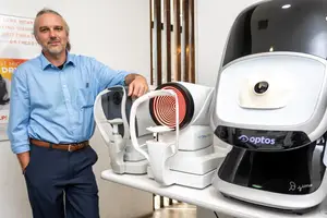 A man is standing next to a white table with three machines on it. The man is wearing a blue shirt and blue pants and is smiling at the camera.