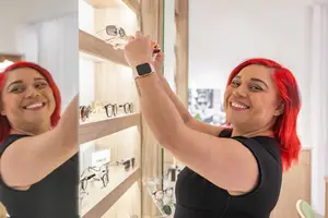 A woman with red hair is smiling and appears to be placing glasses on a shelf while wearing a black sleeveless top and a watch
