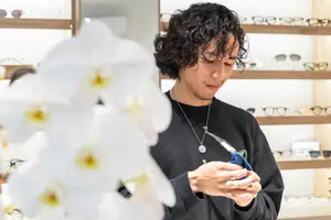 A man wearing a black sweatshirt is adjusting his glasses in front of a white flower. Behind him, shelves with eyeglasses are displayed, and a few are placed on the shelves. The man is looking at something in front of him, and he is holding a blue cloth in his hand. The shelves are arranged in a row, and some are illuminated by a bright light.