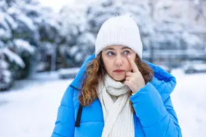 A woman wearing a blue jacket, white beanie, and white scarf is standing in a snowy area and posing for a photo with her right hand on her face.