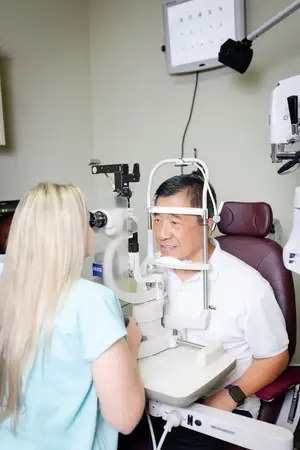 An older man sits in an eye exam chair while a woman in a white shirt adjusts the equipment.