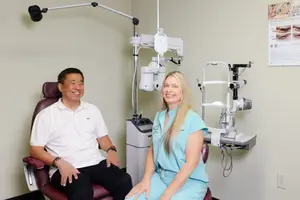 A man and a woman sitting in chairs inside an eye clinic, with a doctor's chair, medical equipment, and a wall with a poster and an outlet in the background.