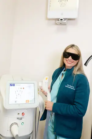 A woman in a blue jacket and sunglasses stands next to a dry eye spa machine and an eye chart on the wall.