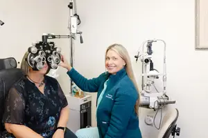 A woman is sitting on a chair while another woman stands beside her, holding a device with the text 'Orth.' near the woman's face.