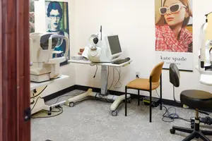 A room with an eye examination machine, two chairs, and a wall with a Kate Spade advertisement poster.
