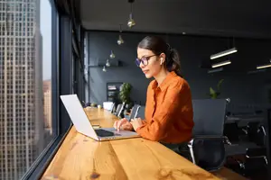 woman in orange shirt wearing glasses sitting at a desk using a laptop