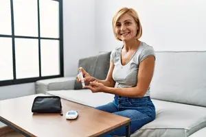 A woman is sitting on a couch in a living room holding a glucometer, smiling, and looking at the camera