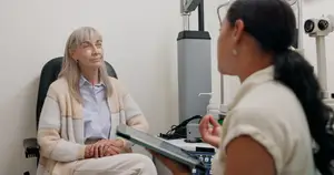 An older woman with glasses is sitting on a chair while a woman in front of her is holding a tablet, probably a doctor in an eye clinic.