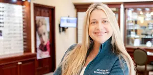 A woman with blonde hair is smiling while standing in a room with a wooden cabinet and a poster on the wall.