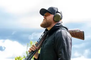 A man with a beard wearing a black jacket and holding a rifle while wearing ear protection and a hat