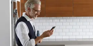 An older man with glasses and a vest stands in a kitchen, looking at his phone with a smile on his face.