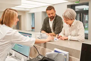 A woman in a white coat is speaking with an elderly woman and a man in a room that appears to be a clinic