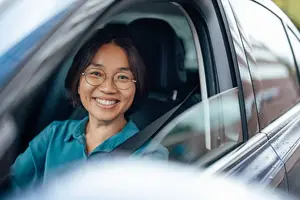 A smiling woman wearing glasses and a blue shirt is sitting in a car with a seatbelt on.
