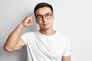 A man wearing glasses and a white t-shirt poses with his hand on his head, possibly posing for a photo